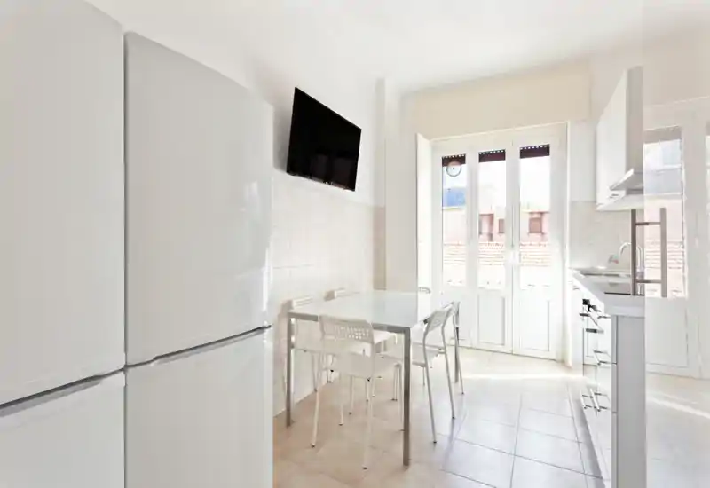 Cucina moderna e luminosa con piccolo tavolo da pranzo, mobili bianchi e grandi porte in vetro che lasciano entrare molta luce naturale — semplice, ariosa e funzionale.
