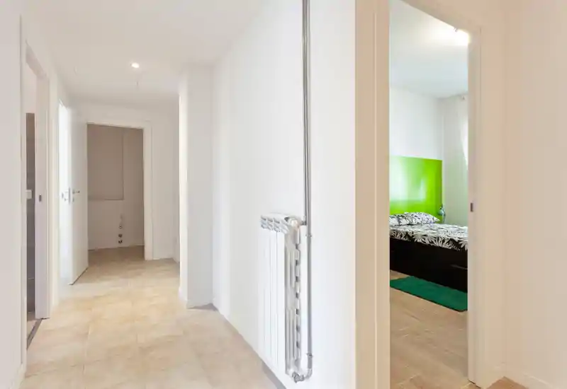 Pasillo luminoso y limpio con suelo de baldosas y radiador a la vista; una puerta a la derecha muestra un vislumbre del dormitorio.