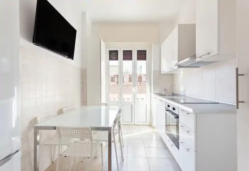 Cocina moderna y luminosa con armarios blancos, horno empotrado y mesa de comedor junto a una puerta de vidrio que aporta abundante luz natural.