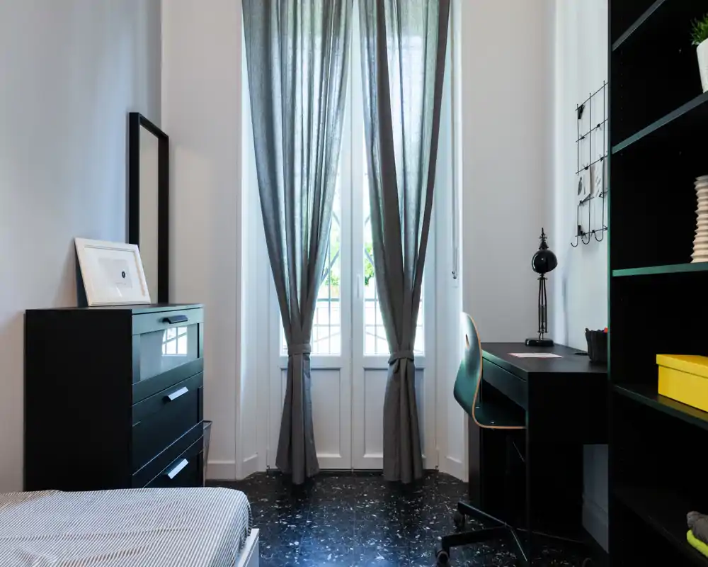 Bright, neatly styled bedroom with a visible single bed, tall window with gray curtains, dresser and a study desk creating a functional yet cozy sleeping area.