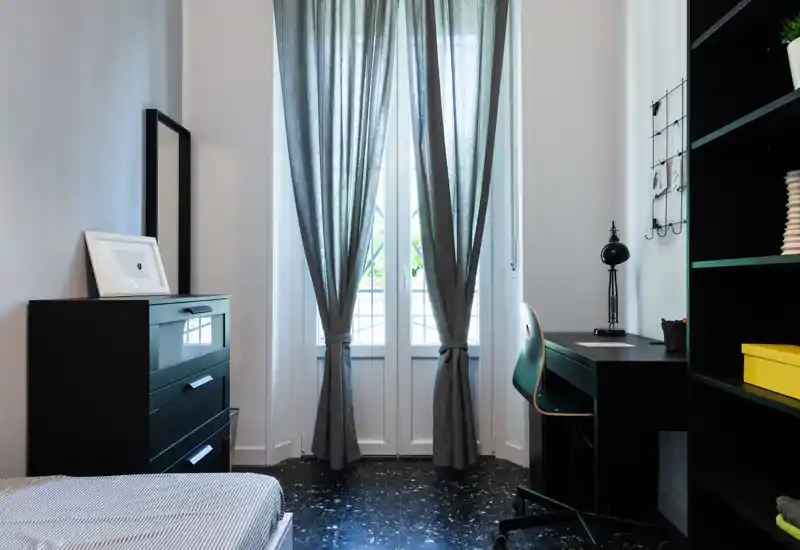 Bright, neatly styled bedroom with a visible single bed, tall window with gray curtains, dresser and a study desk creating a functional yet cozy sleeping area.