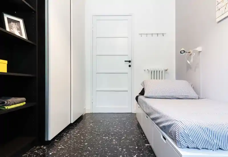 Small, modern single bedroom with a neatly made bed, built-in storage and a minimalist reading lamp. The room features striped bedding, a black shelving unit and a clean white door, ideal for a compact rental.