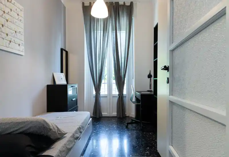 Bright, tidy bedroom with a single bed, desk and natural light from tall windows; neutral décor and practical storage make it ideal for a student or short-term rental.