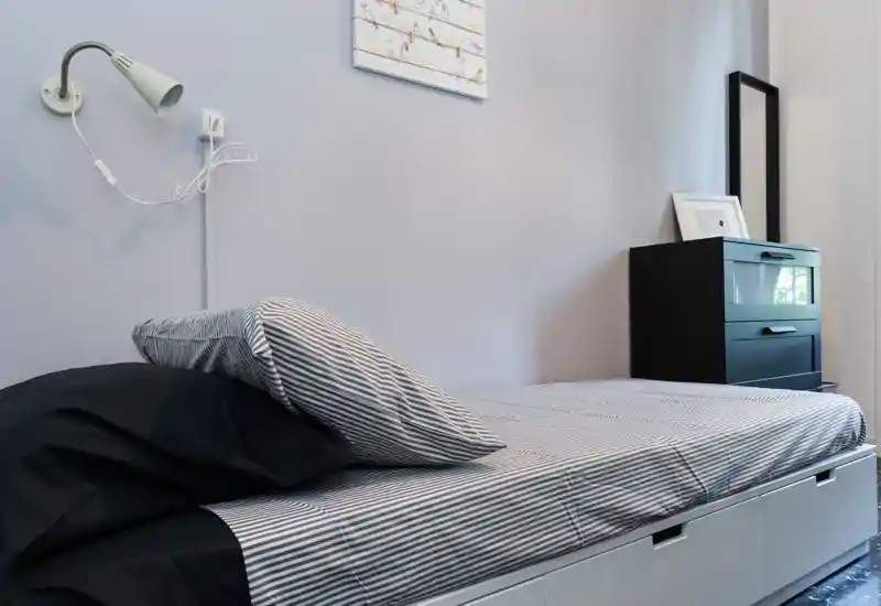 Simple, modern single bedroom with a striped fitted sheet, pillows and under-bed storage drawers. Soft wall lamp and a compact chest of drawers make this a practical, tidy sleeping area.