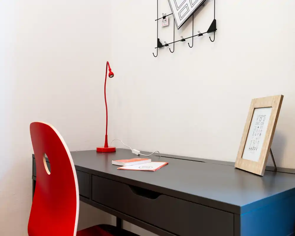 Zona scrivania luminosa e minimal con tavolo da lavoro nero, sedia rossa e lampada rossa abbinata; accessori ordinati come una stampa incorniciata e un quaderno — perfetta per lavorare o studiare.