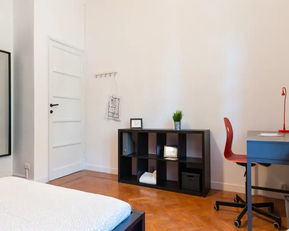 Angolo camera luminoso e minimal con angolo di letto visibile, una libreria bassa e una piccola scrivania: linee pulite e pavimento in legno creano un'atmosfera calma.