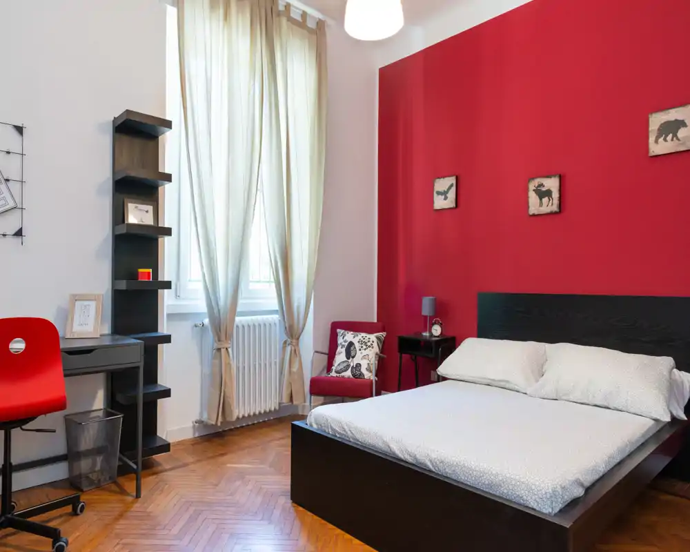 Camera da letto luminosa e di design con letto matrimoniale, parete rossa a contrasto e luce naturale da una grande finestra; include una scrivania e una poltrona per la lettura.