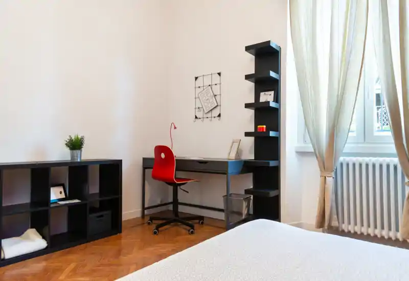 Camera da letto luminosa e ordinata con letto in primo piano, scrivania funzionale e scaffalature, illuminata da una grande finestra.