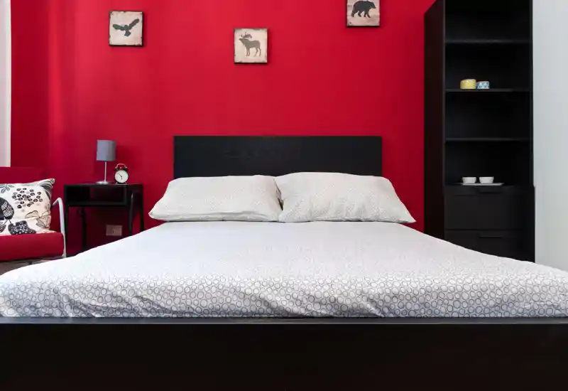 Camera da letto elegante con un ampio letto matrimoniale al centro su una parete rossa; biancheria pulita, comodino con lampada e sveglia e mensole che creano un ambiente ordinato e accogliente.