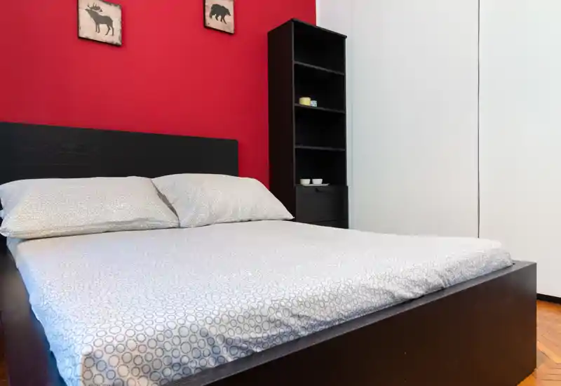 Camera da letto luminosa e ben arredata con struttura del letto in legno scuro su una parete rossa, biancheria a motivo e una alta libreria: pulita, accogliente e pronta per le foto.