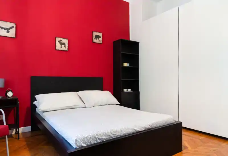 Camera da letto luminosa e ordinata con letto matrimoniale davanti a una parete accentuata rossa, scaffalatura minimale e grande armadio bianco — perfetta per un soggiorno accogliente in città.