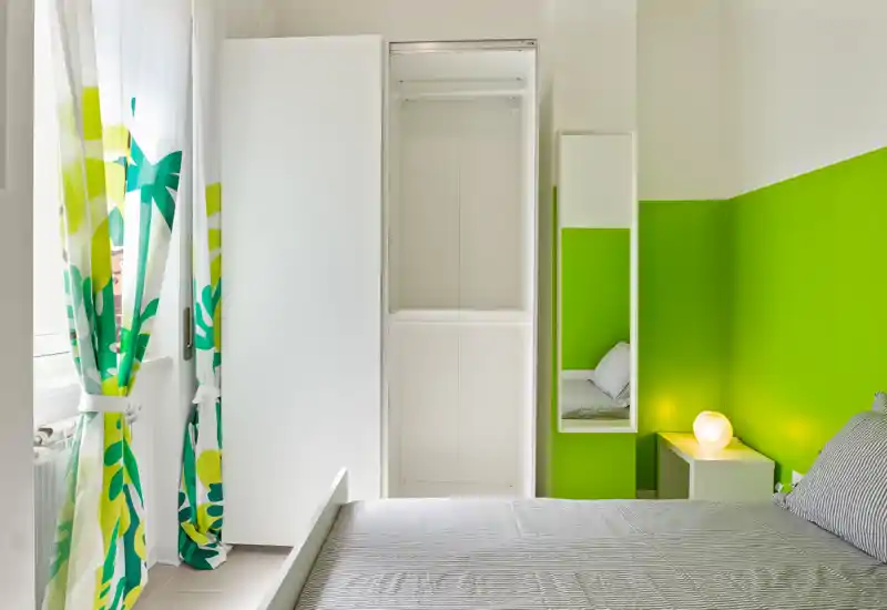 Dormitorio moderno y luminoso con cama gris, pared verde como acento y cortinas estampadas; mesita con lámpara y armario alto blanco para almacenamiento.