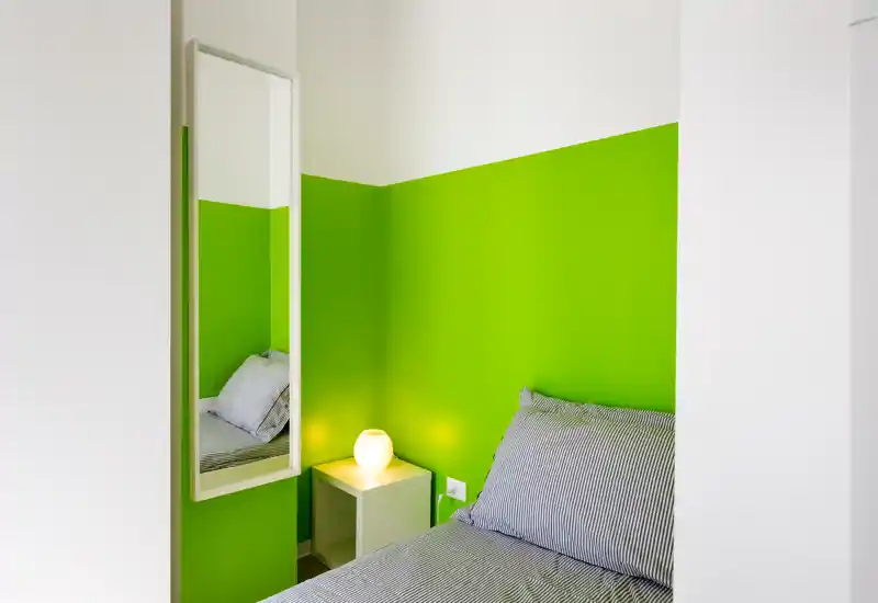 Rincón de dormitorio pequeño y luminoso con una cama individual bien hecha junto a una pared verde intensa, mesita con lámpara cálida y un espejo alto que refleja parte de la cama.