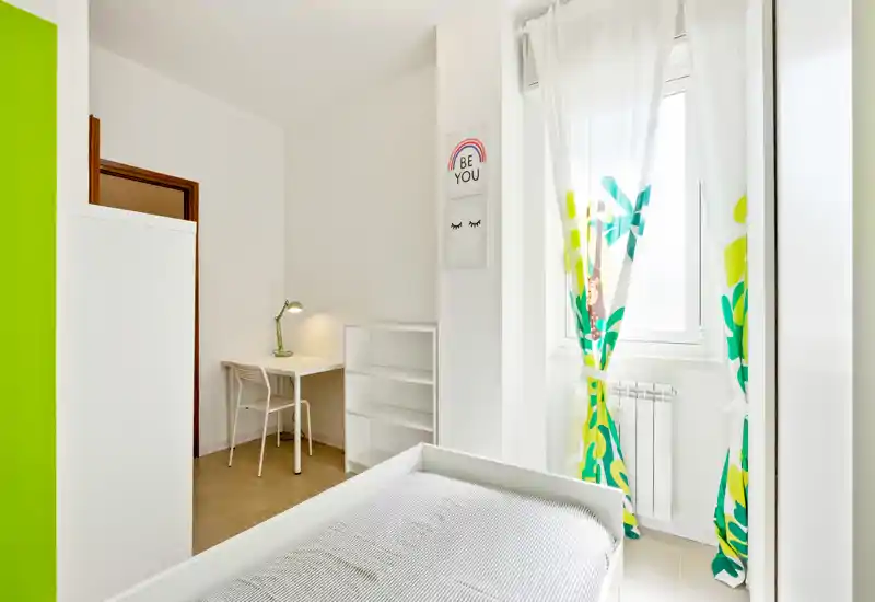 Dormitorio pequeño y luminoso con cama individual, escritorio y estantería. Paredes blancas y cortinas coloridas que dan una sensación fresca y acogedora, ideal para estudiantes o una sola persona.