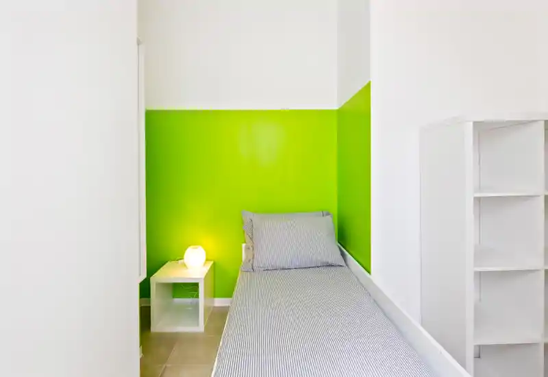 Habitación individual compacta con cama bien hecha, ropa de cama a rayas, mesita con lámpara y una pared verde llamativa; luminosa, minimalista y bien preparada para alquileres.