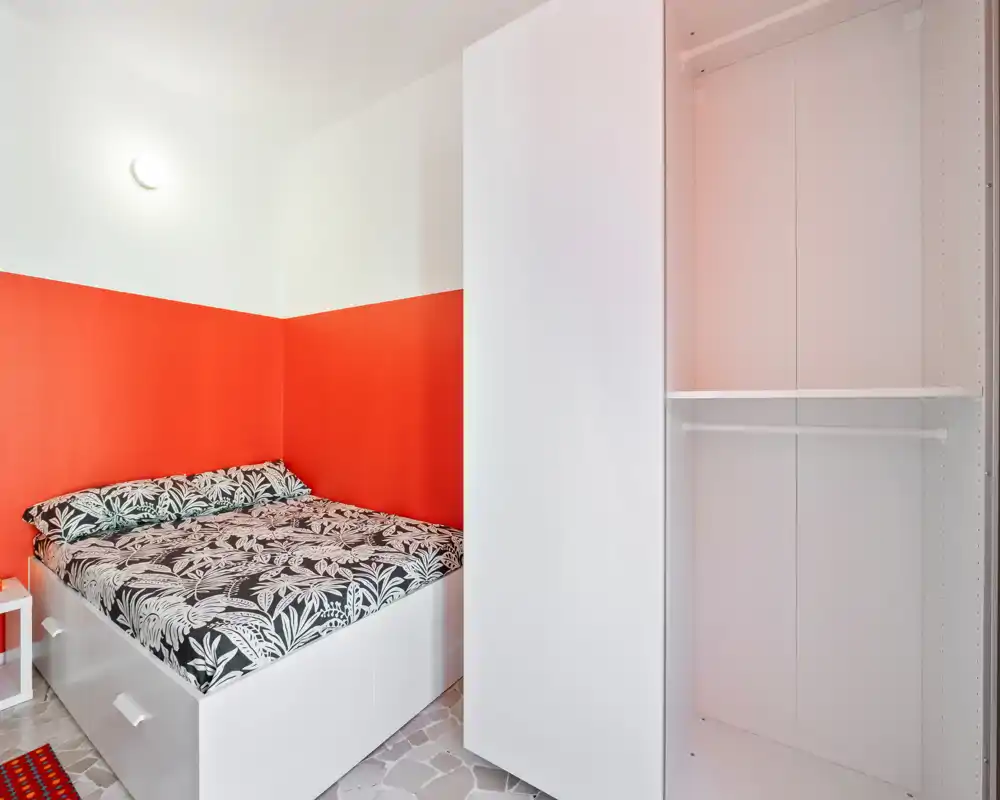 Dormitorio compacto y luminoso con cama doble, ropa de cama estampada y armario blanco empotrado. La pared naranja y la distribución ordenada generan un ambiente moderno y acogedor.