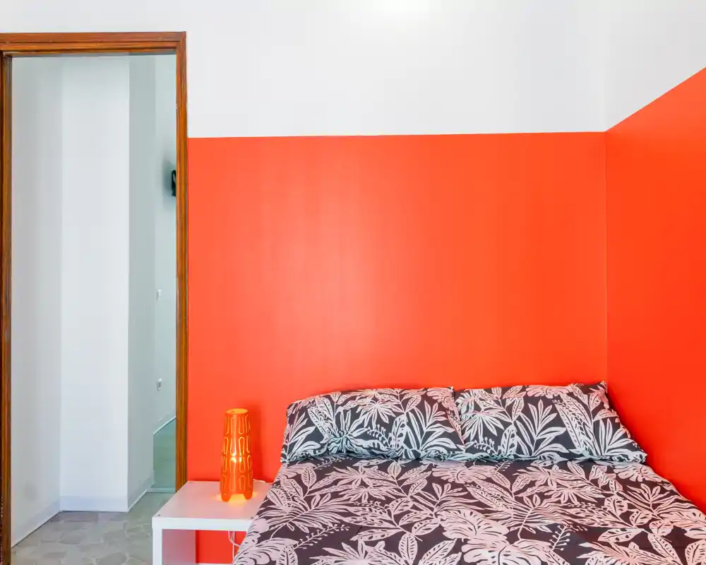 Dormitorio moderno y luminoso con cama doble estampada frente a una pared roja intensa, mesita y lámpara; estilo limpio y minimalista, perfecto para anuncios.