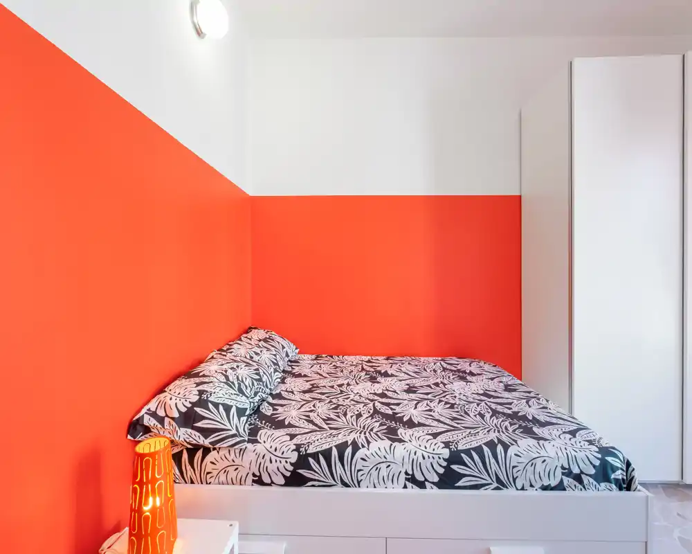 Dormitorio luminoso y bien presentado con cama de matrimonio y ropa de cama estampada, paredes coral llamativas y armario blanco. La habitación se ve ordenada, moderna y acogedora, perfecta para un anuncio de alquiler.
