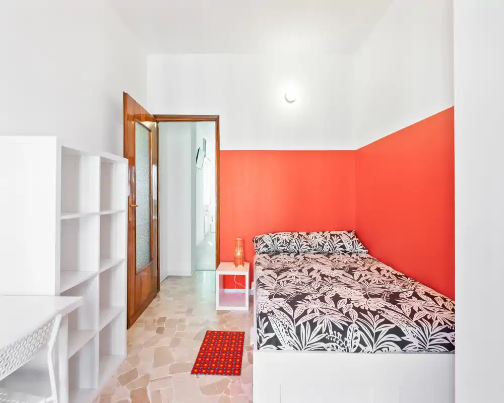 Dormitorio luminoso y moderno con cama doble estampada frente a una pared roja, mesita con lámpara y estantería blanca que ofrece almacenamiento y espacio de trabajo.