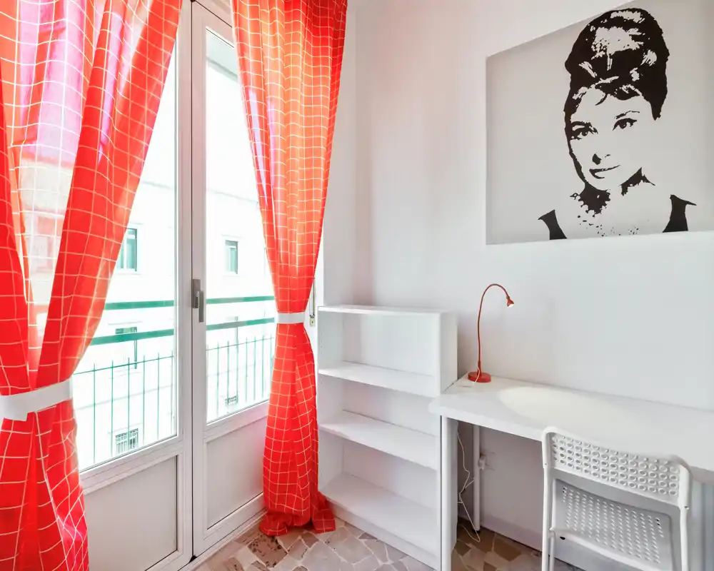 Rincón de trabajo luminoso con escritorio blanco, silla y estantería junto a una gran ventana/puerta con cortinas rojas. Estilo minimalista y limpio con cuadro decorativo y lámpara de escritorio, ideal para concentrarse.