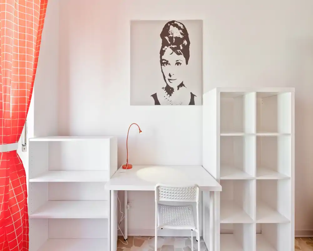 Rincón de trabajo luminoso y minimalista con escritorio blanco, silla y estanterías abiertas junto a una ventana con cortinas rojas — un espacio ordenado ideal para teletrabajo o lectura.