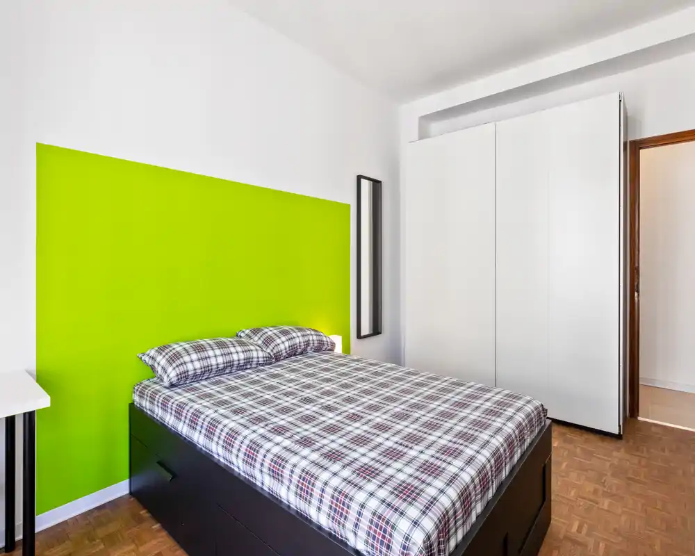 Dormitorio moderno y luminoso con cama doble, ropa de cama a cuadros y una pared verde llamativa; líneas limpias y un gran armario blanco que aportan un estilo minimalista.