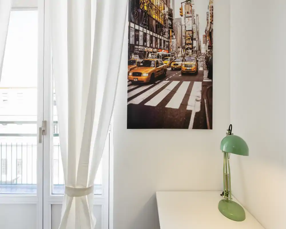 Rincón luminoso de la sala con gran ventana, cortinas claras, cuadro urbano y una lámpara de mesa — aireado, minimalista y bien iluminado.