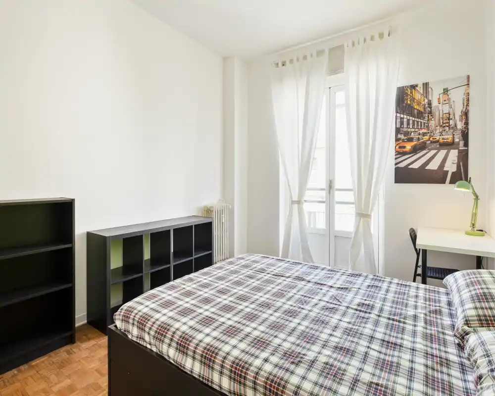 Dormitorio luminoso y bien decorado con cama doble de estampado a cuadros, estanterías, escritorio y una gran ventana con cortinas translúcidas que dejan entrar la luz natural.