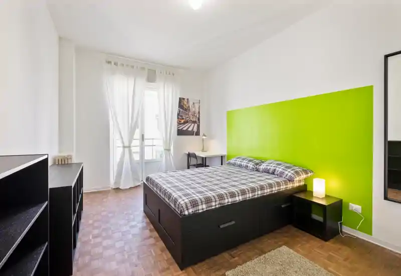 Dormitorio luminoso y bien decorado con cama doble, mesita y escritorio; gran ventana con cortinas traslúcidas que aporta abundante luz natural y sensación moderna.