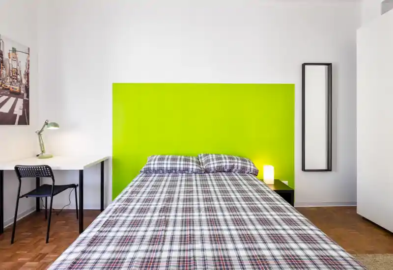 Dormitorio moderno y luminoso con cama centrada frente a un cabecero verde brillante, lámpara de mesita y un escritorio sencillo. Líneas limpias y fuerte contraste cromático crean un espacio acogedor y bien iluminado.