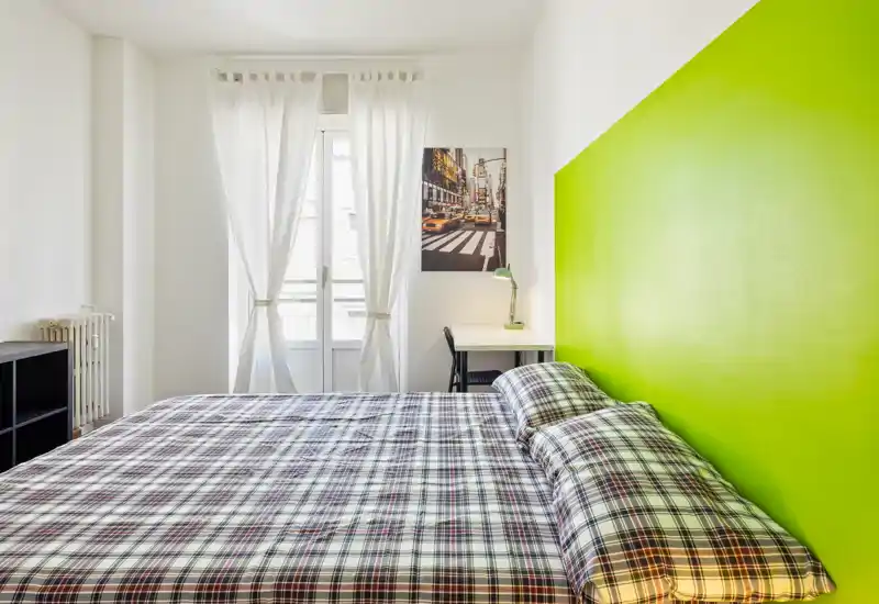Dormitorio moderno y luminoso con una gran cama doble frente a la ventana y una pared verde de acento; la ropa de cama a cuadros y un pequeño escritorio hacen el espacio práctico y acogedor.