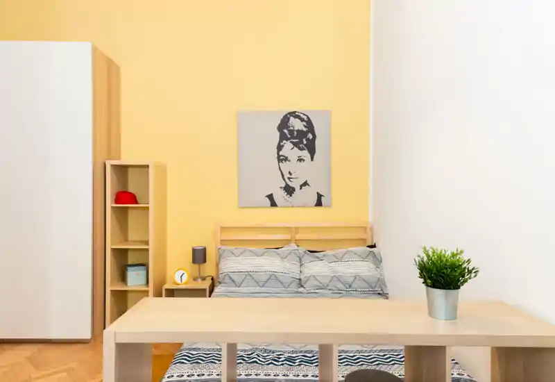 Dormitorio luminoso y bien decorado con cama de madera, ropa de cama con estampado y pared amarilla. Almacenamiento minimalista, lámpara de mesita y cuadro decorativo crean un espacio acogedor y atractivo para anuncios.