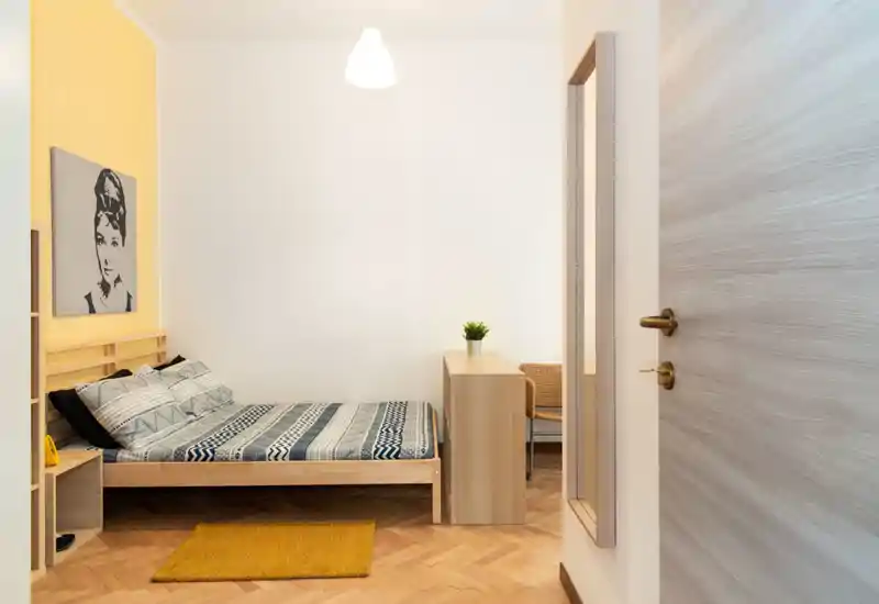 Dormitorio luminoso y ordenado con cama de madera, ropa de cama con estampado y acentos amarillos cálidos. La habitación tiene un escritorio pequeño, un cuadro en la pared y una alfombra acogedora.