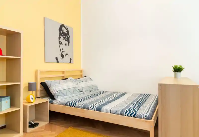 Dormitorio luminoso y de estilo sencillo con estructura de cama de madera, ropa de cama estampada y muebles de madera clara; pared amarilla y detalles mínimos crean un ambiente acogedor y moderno.