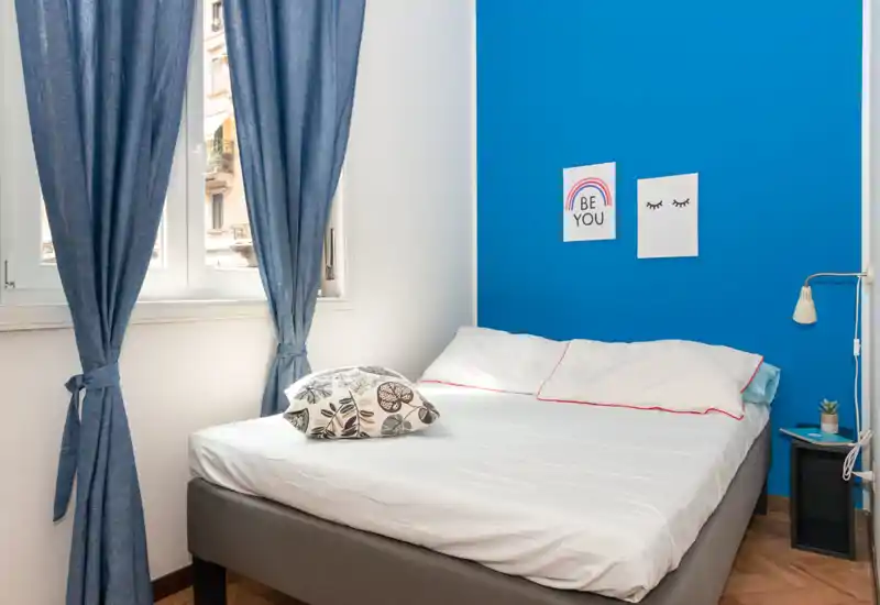 Dormitorio luminoso y bien arreglado con cama doble contra una pared azul intensa, luz natural desde la ventana y decoración de mesita sencilla.