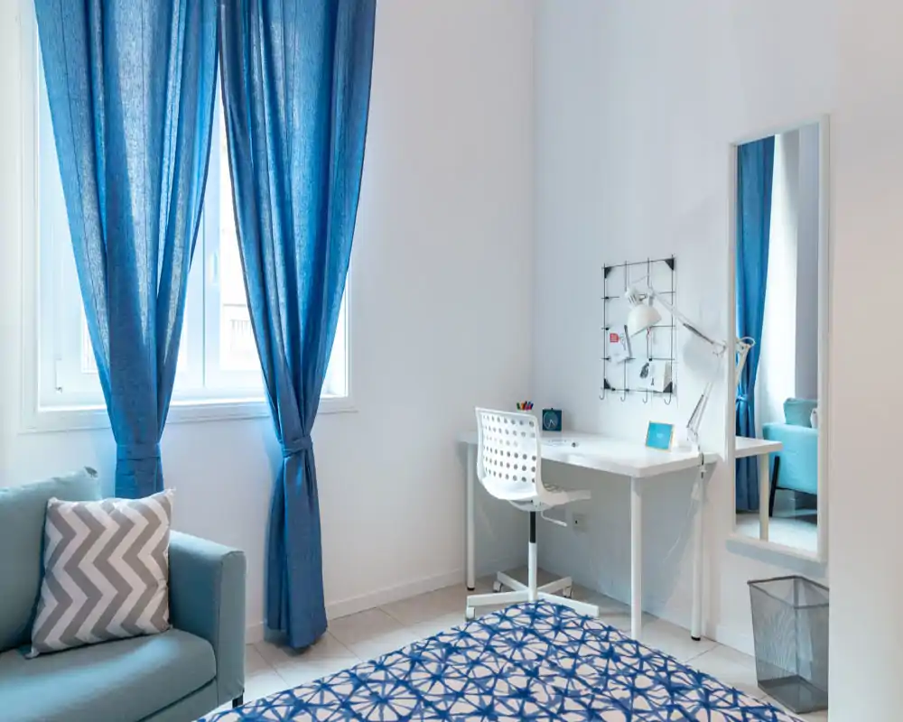 Rincón de dormitorio luminoso y moderno con cortinas azules, colcha estampada y un escritorio blanco ordenado — ideal para invitados o estudiantes. El espacio se percibe fresco y bien iluminado con textiles coordinados y mobiliario funcional y minimalista.