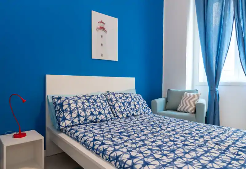Dormitorio moderno y luminoso con cama de estructura blanca, ropa de cama azul estampada y un sillón junto a la ventana; estilo fresco con buena luz natural.
