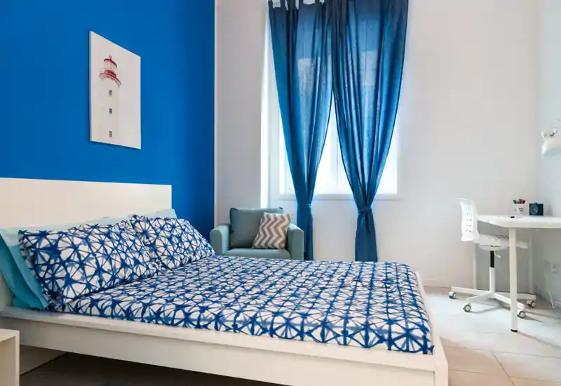 Dormitorio moderno y luminoso con cama doble grande estampada, pared azul de acento, un sillón y una zona de trabajo con escritorio y silla junto a la ventana.