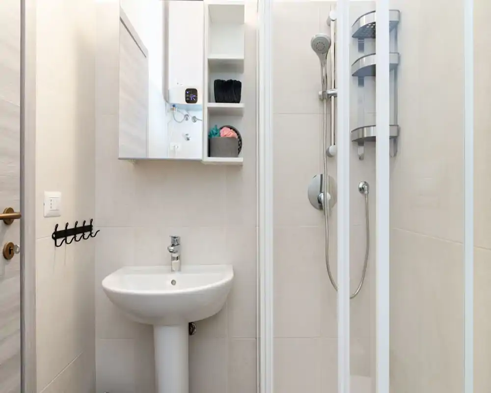 Bagno moderno e compatto con lavabo a colonna, specchio con mobiletto e doccia con pareti in vetro e soffione doccia estraibile. Piastrelle chiare e ripostiglio ordinato rendono l’ambiente funzionale e pulito.