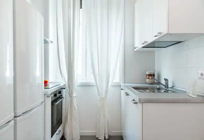 Cocina moderna y compacta, luminosa, con armarios blancos, fregadero de acero inoxidable y ventana con cortinas claras que aportan sensación de frescura.