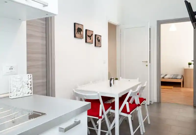 Rincón de comedor moderno y luminoso con mesa blanca y sillas con cojines rojos, junto a una cocina compacta y la entrada a un dormitorio.