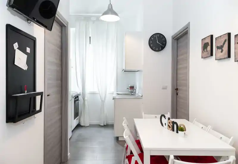 Zona pranzo moderna e luminosa adiacente a una cucina compatta. Tavolo bianco con sedie pieghevoli, quadri decorativi e luce naturale da una finestra con tenda rendono l'ambiente accogliente.