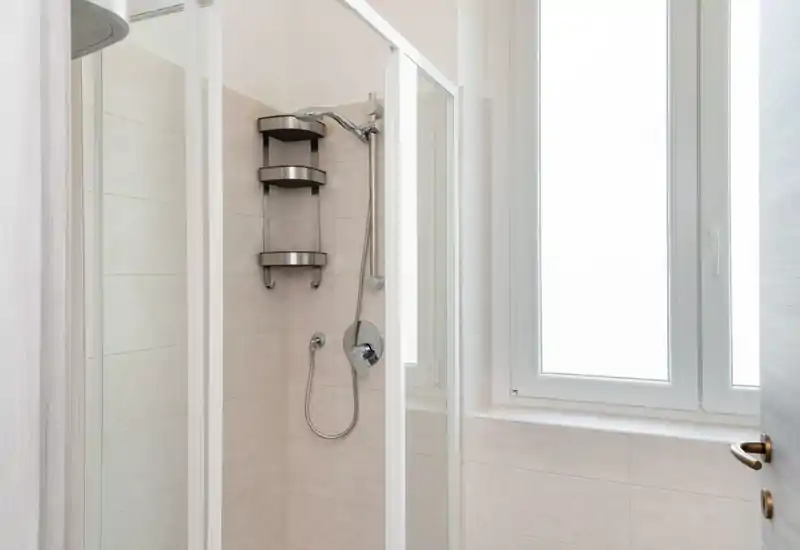 Rincón de baño moderno y luminoso con mampara de ducha de cristal, cabezal cromado y paredes alicatadas junto a una gran ventana esmerilada.