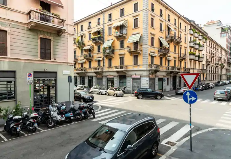 Esquina de calle urbana soleada con coches y motos aparcados frente a edificios clásicos — ideal para mostrar la ubicación y el ambiente del barrio.