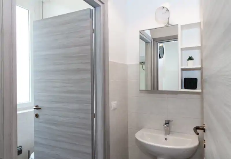 Baño pequeño y moderno con lavabo mural, armario con espejo, azulejos grises neutros y luz natural suave desde una ventana cercana.