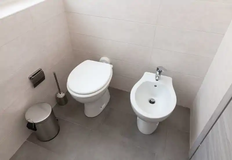 Baño compacto y moderno con inodoro y bidé sobre suelo de baldosas, accesorios de acero inoxidable y azulejos de pared de tonos neutros.