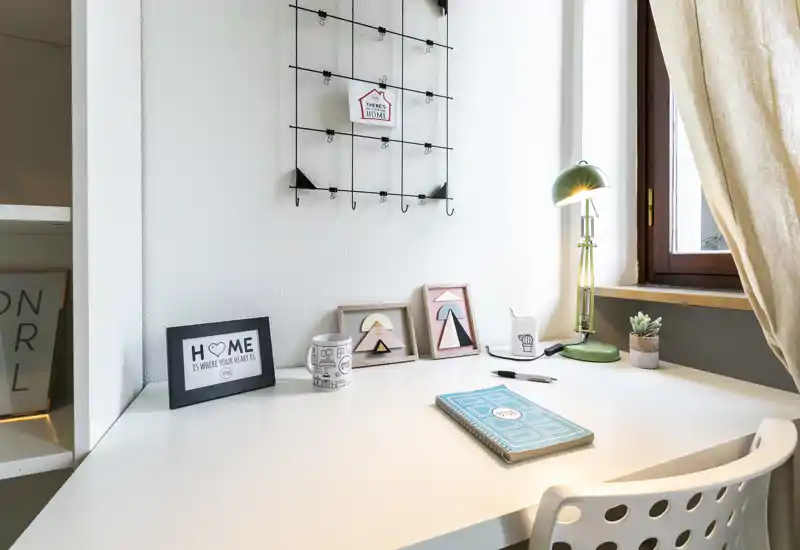 Angolo scrivania luminoso e arredato con cornici decorative, una lampada verde e una piantina — ideale come postazione studio vicino alla finestra.