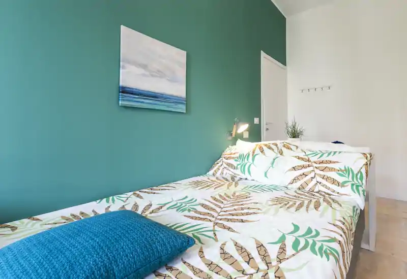 Bright, styled bedroom with a double bed against a teal accent wall, patterned bedding and a bedside lamp creating a cozy, modern feel.