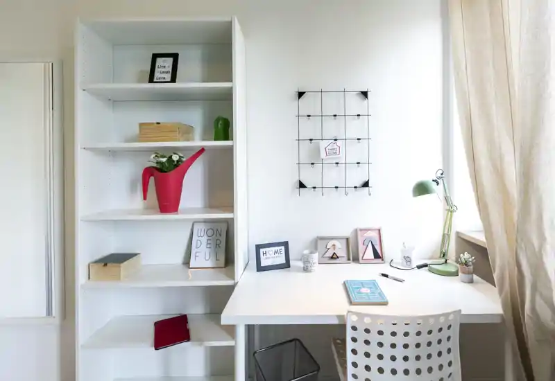 Angolo lavoro luminoso e ordinato con scrivania bianca, sedia forata, scaffale e dettagli decorativi — ideale per lavorare o leggere alla luce naturale della finestra.