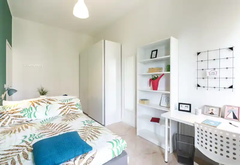 Bright, modern bedroom with a patterned double bed in the foreground, a tall white wardrobe and shelving, and a small desk by the window — fresh, well-lit and neatly styled for short-term rental photos.